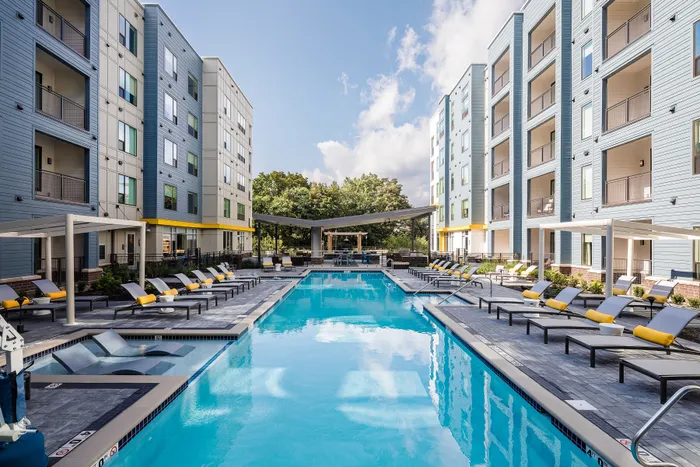 One-Bedroom Luxury Apartments In Strip District, PA - Edge 1909 - A Swimming Pool Surrounded By Grey Pavers, Modern Lounge Chairs With Yellow Pillows, And Multi-Story Blue And Tan Apartment Buildings.