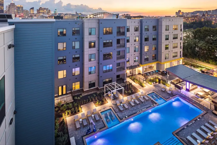Modern apartment complex at sunset with a shimmering swimming pool, stylish outdoor seating, and vibrant city skyline views.