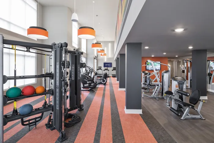 Strip District, PA, Luxury Studio Apartments - Edge 1909 - A Modern Fitness Center With Orange-Striped Functional Turf, Hoist Strength Machines, And Orange Drum Pendant Lighting.