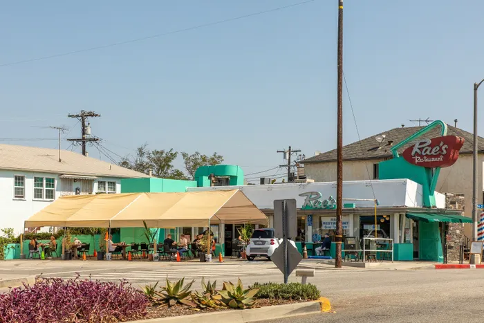 Charming roadside diner with teal and white facade, sheltered outdoor seating, and thriving desert plants, captures vintage Americana style.