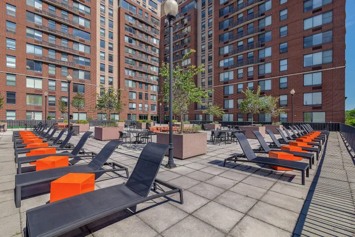 Luxurious apartment complex with sunlit outdoor patio featuring stylish lounge chairs, vibrant orange accents, and lush greenery.