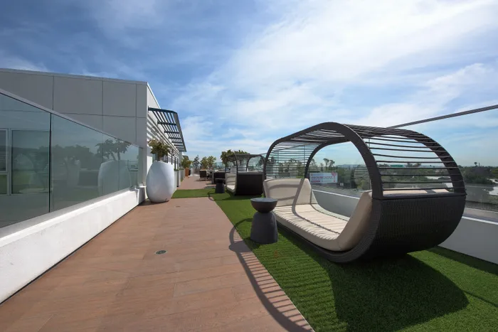 Bright anPet-Friendly Luxury Apartments in Sawtelle, CA - C on Pico - Rooftop Terrace with a Row of Chic Daybeds, Side Table, Green Turf, Planters, and Glass Railings Providing Expansive City Views.d stylish rooftop terrace featuring chic lounge furniture, lush greenery, and expansive city views perfect for relaxation.
