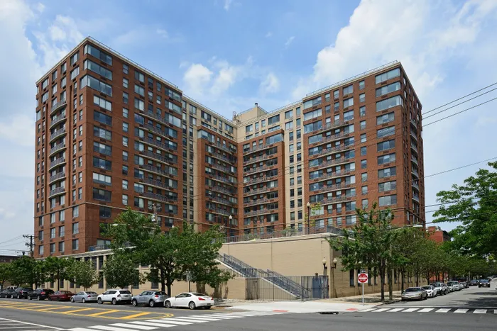 3-Bedroom Luxury Apartments In Hoboken, NJ - 77 Park Avenue - A Large L-Shaped Brick Apartment Building With Many Balconies On A City Corner Under A Cloudy Sky.