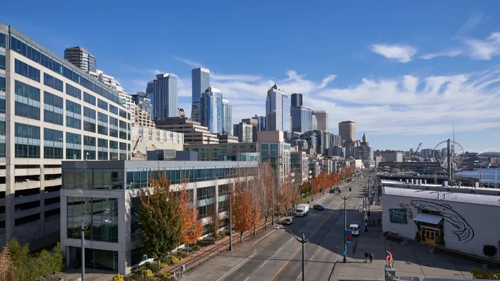 Scenic urban skyline with modern architecture, vibrant autumn trees, and waterfront in the foreground, showcasing city life with a backdrop of clear blue skies.