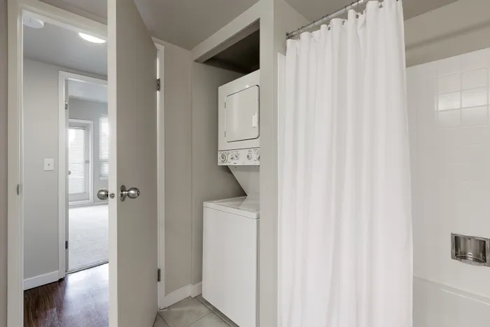 Modern apartment bathroom with in-unit stacked washer and dryer, white tiled shower, and bright wooden flooring leading to an airy living space.