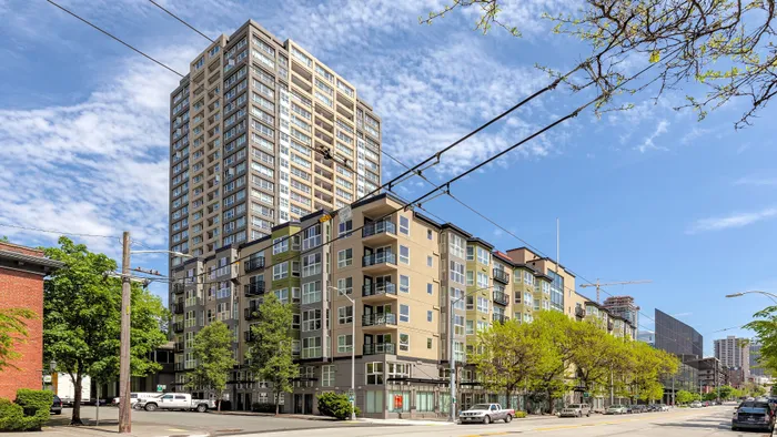Modern high-rise apartment building with large windows, balconies, and a vibrant cityscape backdrop, under a bright blue sky.