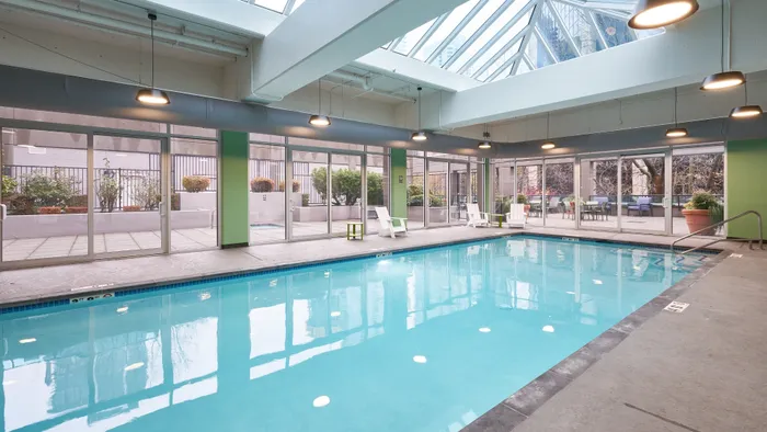 Luxurious indoor pool with skylight ceiling, surrounded by modern lounge chairs and landscaped patio views.