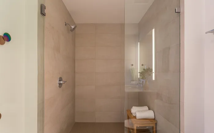 Luxurious modern shower with elegant beige tiles and a sleek glass door, complemented by a cozy wooden bench with fresh towels and a warm ambiance.