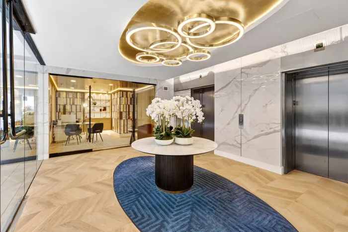 Luxurious modern lobby with elegant marble walls, sleek elevators, and a stunning circular light fixture, complemented by a stylish blue carpet and flourishing floral centerpiece.