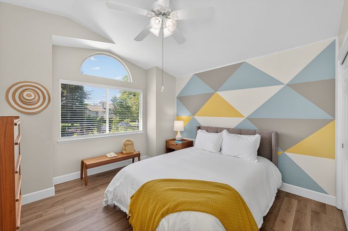 Bright and inviting bedroom featuring a modern geometric accent wall, cozy bed with a mustard throw, and a large window offering serene garden views.