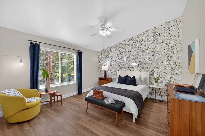 Inviting bedroom with stylish decor, featuring a patterned accent wall, hardwood floors, and cozy sitting area with a mustard armchair and natural light.