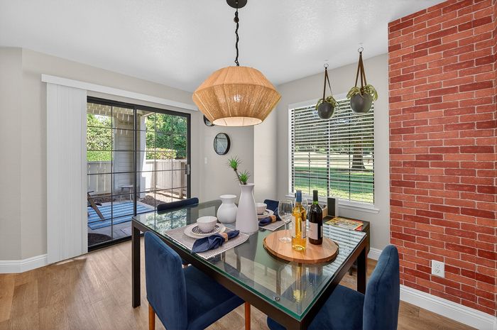 Elegant dining room with chic glass table, modern pendant light, and a stunning red brick accent wall, overlooking a lush garden patio through large sliding doors.