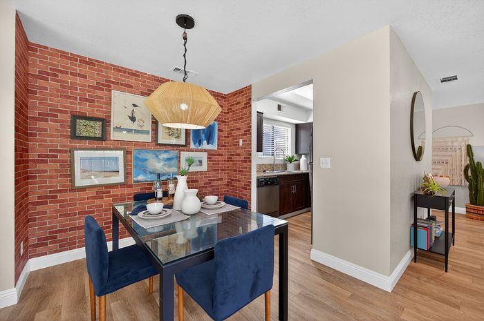 Chic dining area with brick accent wall, modern glass table, and plush blue chairs leading to an open kitchen space.