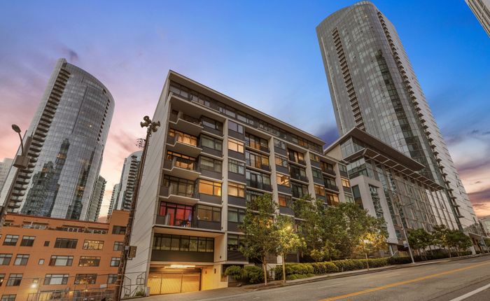 Modern urban apartment building with sleek architecture and large windows, surrounded by tall skyscrapers at sunset.