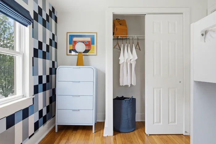 Bright and modern bedroom corner featuring a sleek white dresser, open closet with organized hanging clothes, and artistic wall decor. Elegant hardwood floors add warmth and charm.