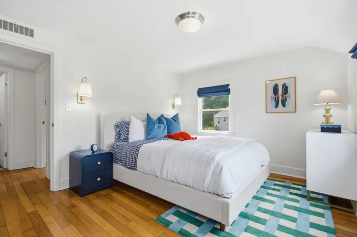 Bright and cozy bedroom featuring elegant hardwood floors, a plush white bed with vibrant accents, and a charming view from the window.