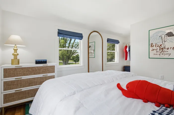 Bright and cozy bedroom with elegant decor, featuring dual windows offering scenic views, a stylish dresser, and a charming stuffed toy on the neatly made bed.
