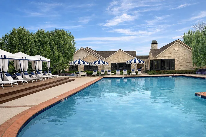 Luxurious outdoor pool area with elegant cabanas and chic blue-and-white umbrellas, surrounded by lush greenery and a beautiful stone clubhouse.