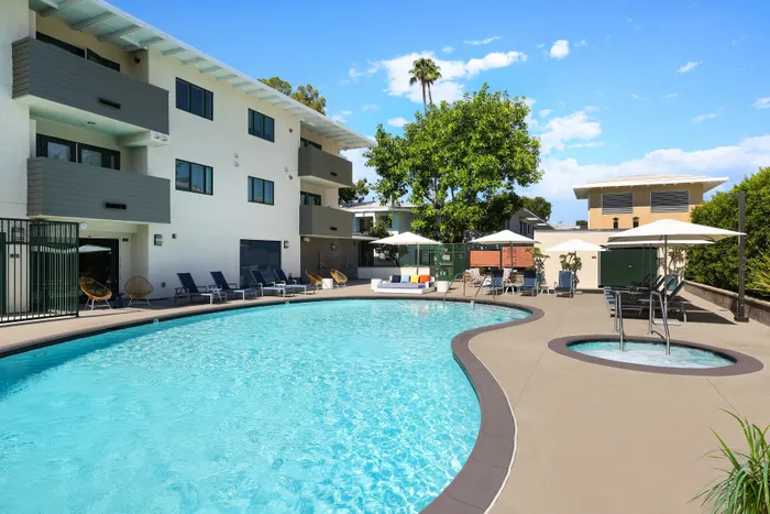 Inviting outdoor pool area with sun loungers and umbrellas, set against a modern apartment building and lush greenery.