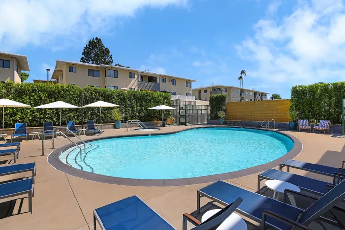 Inviting apartment community featuring a sparkling pool, surrounded by stylish lounge chairs and elegant umbrellas under a clear blue sky.