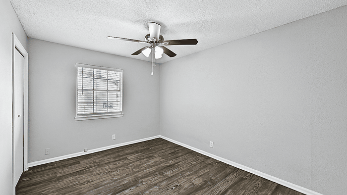 Bright empty room with elegant ceiling fan and large window, featuring rich wooden flooring and neutral wall tones.