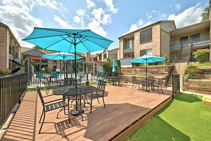 Charming apartment complex featuring a modern outdoor patio with turquoise umbrellas, stylish seating, and a refreshing pool in the background.