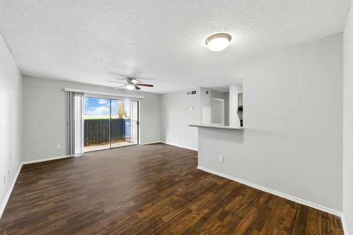 Contemporary living room with sleek hardwood floors, a ceiling fan, and a bright balcony view, seamlessly connected to an inviting open kitchen.