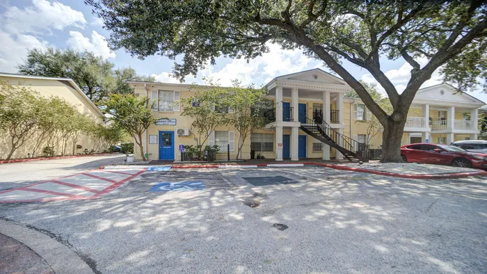 Charming apartment complex with classic architectural style, featuring a lush tree-lined entrance, spacious parking area, and inviting leasing office with vibrant blue accents.