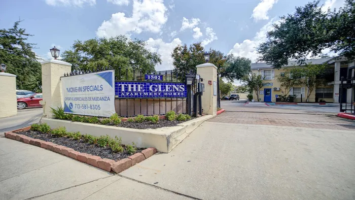 Inviting entrance to The Glens Apartment Homes featuring a sign with move-in specials, surrounded by lush greenery and bright skies.