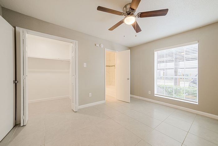 Bright and airy bedroom with sleek floor tiles, a ceiling fan, and a large window, featuring an open closet and adjacent modern bathroom.