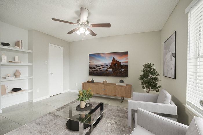 Modern living room with elegant decor including a ceiling fan, sleek furniture, and a large wall-mounted TV showcasing coastal imagery. Features built-in shelves and abundant natural light.