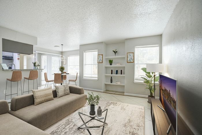 Bright and modern living room with cozy seating, sleek decor, and abundant natural light, open to a stylish kitchen and dining area with elegant bar stools and lush greenery.