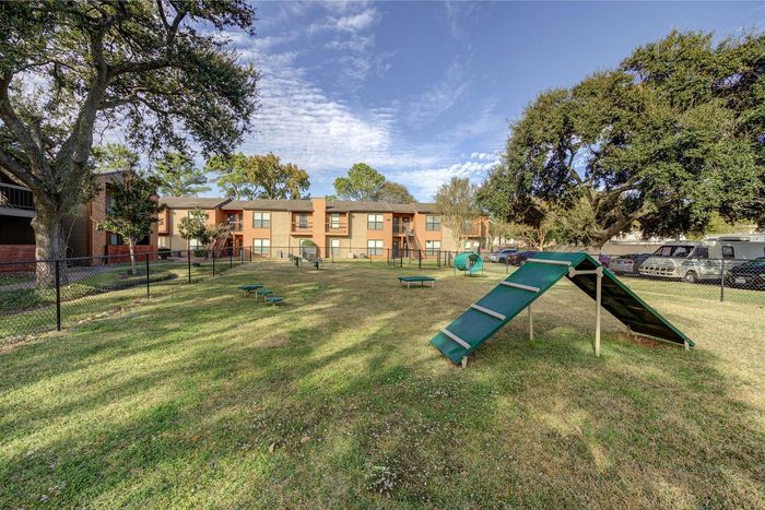 Inviting apartment complex with lush green lawns, a pet-friendly play area, and majestic trees under a clear blue sky.