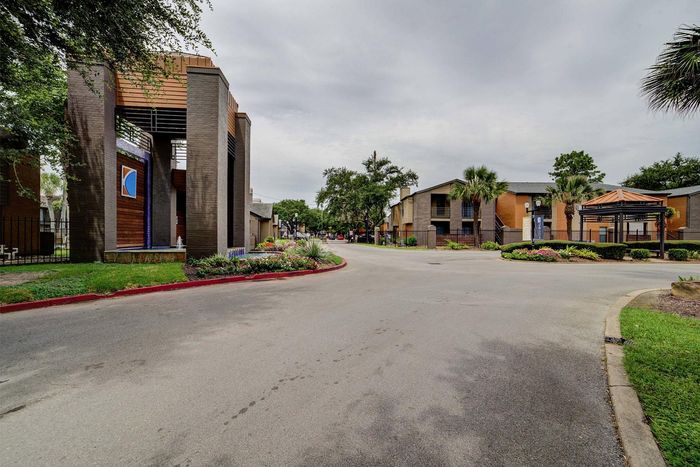 Charming apartment complex entrance surrounded by lush greenery, showcasing modern architecture with palm trees and vibrant garden landscaping.