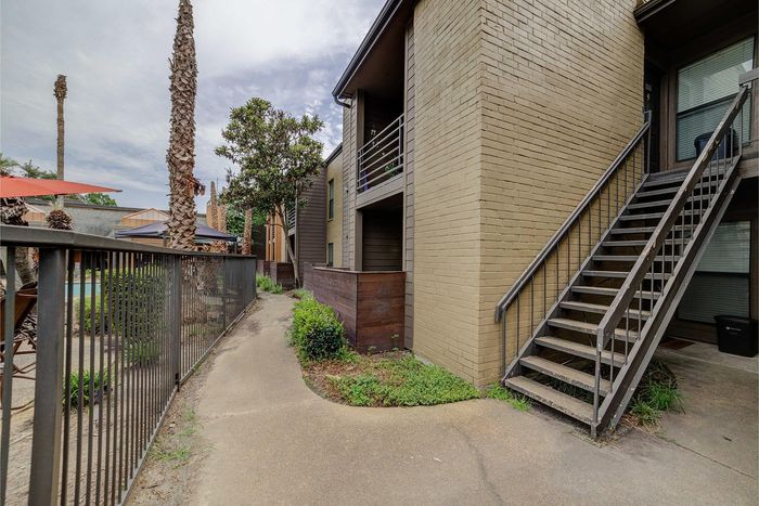 Inviting apartment entrance featuring exterior stairs and lush greenery, next to a fenced pathway leading to a pool area with umbrellas.