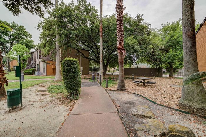 Charming apartment complex exterior with lush greenery, featuring a serene pathway, picnic area, and towering trees for a peaceful outdoor setting.
