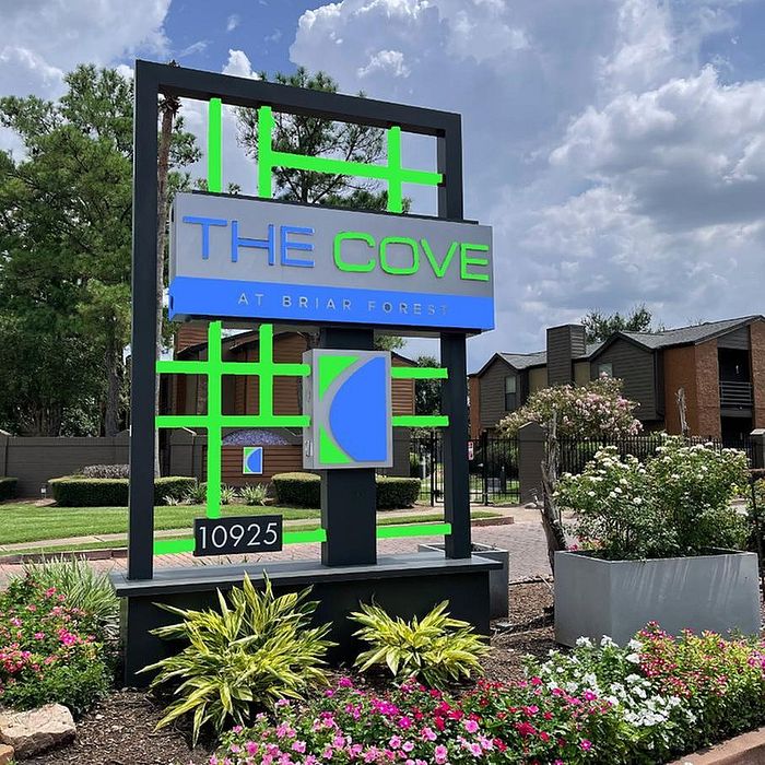 Inviting entrance sign for The Cove at Briar Forest, surrounded by lush greenery and vibrant flowers, under a bright blue sky.