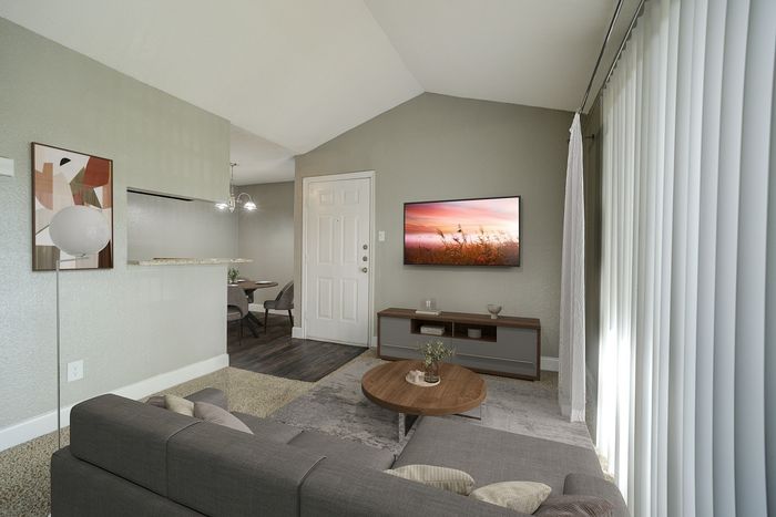 Elegant living room with modern decor, featuring a cozy grey sofa, artistic wall art, mounted TV, and a stylish coffee table. Sunlight streams through vertical blinds, illuminating the tasteful open-plan design with a glimpse of the dining area and chandelier.