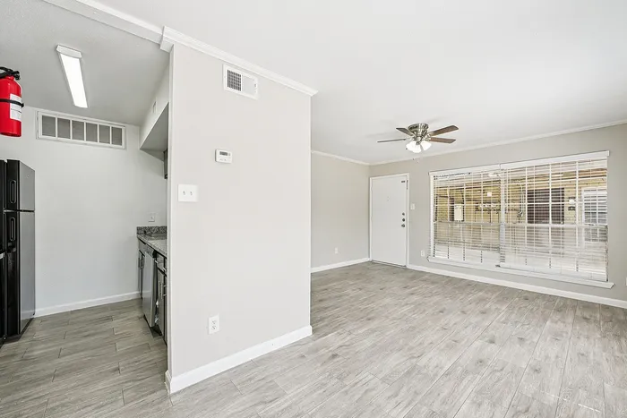Bright and inviting living area with modern gray flooring, ceiling fan, and large windows offering ample natural light, adjacent to a sleek kitchen with dark appliances.