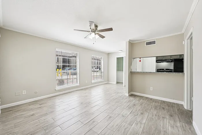 Bright and airy living room with stylish wood-look flooring, large windows for natural light, and a sleek ceiling fan, adjacent to a cozy kitchen area.