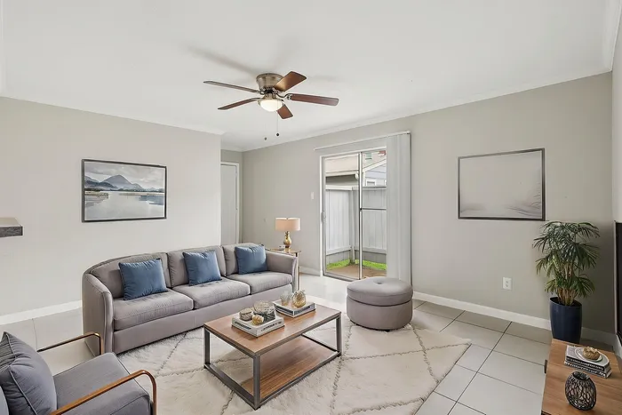 Bright and inviting living room with elegant gray sofa, plush cushions, modern coffee table, and sliding glass doors opening to a private patio. Contemporary decor with ceiling fan and art adorns the walls, creating a cozy ambiance.