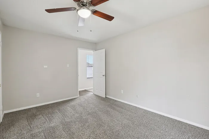 Bright bedroom with plush carpet, ceiling fan, and natural light through a window, offering a cozy and inviting space.