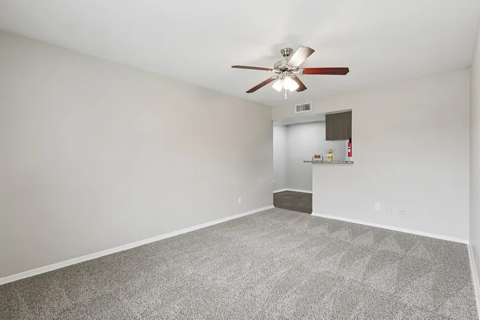 Modern living area with plush carpet, ceiling fan, and open kitchen access.