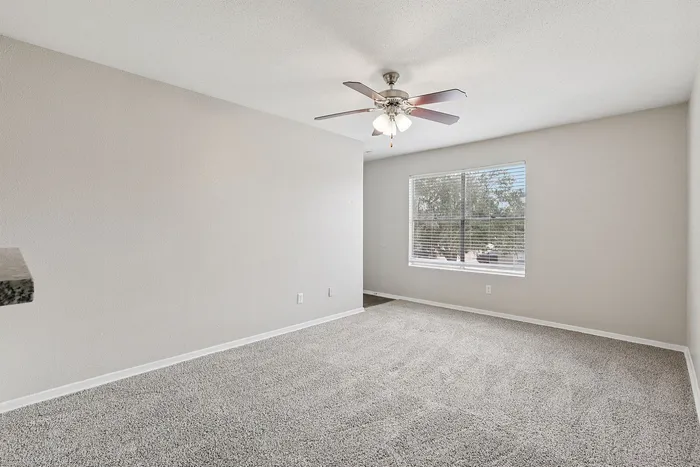 Bright and spacious carpeted living area with a large window and ceiling fan, perfect for relaxation and comfort.