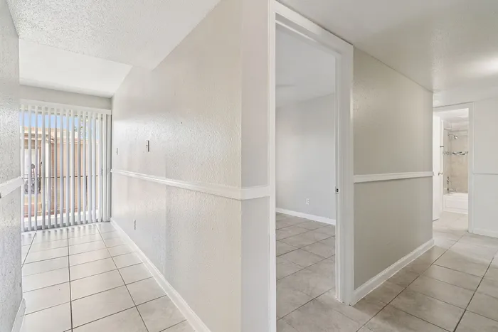 Bright hallway with sleek tile flooring, leading to multiple rooms and a modern bathroom, accentuated by large vertical blinds for natural light.