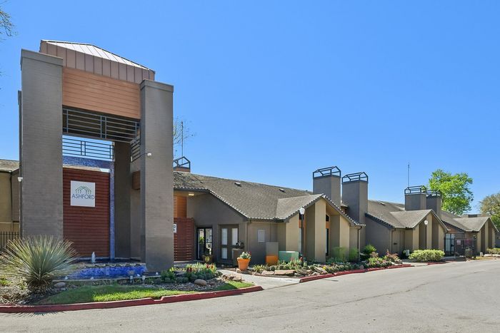 Charming apartment complex with a modern design and welcoming entrance, accented by lush landscaping and a bright, clear sky.