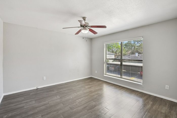Bright and spacious room featuring elegant wood flooring, large window with blinds, and a stylish ceiling fan.