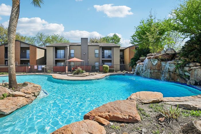 Inviting apartment complex with a sparkling swimming pool, stunning waterfall feature, and lush green landscaping under a clear blue sky.