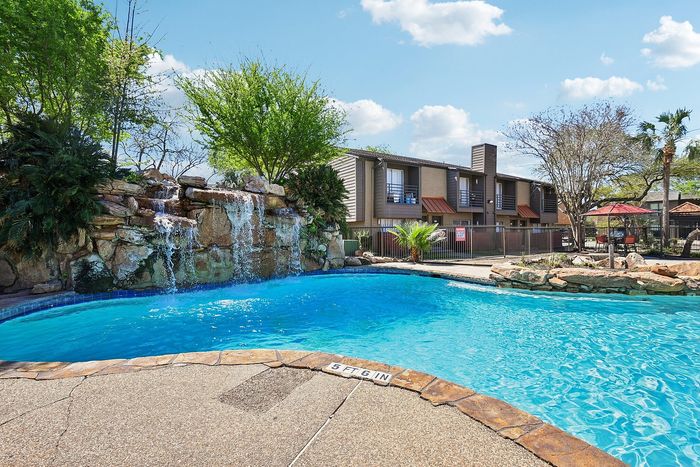 Inviting apartment pool area with a cascading waterfall feature, surrounded by lush greenery and modern buildings under a bright blue sky.