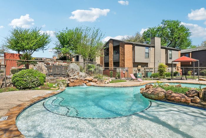 Inviting residential complex boasting a serene outdoor pool with natural rock landscaping, surrounded by lush greenery and modern apartment buildings under a clear blue sky.
